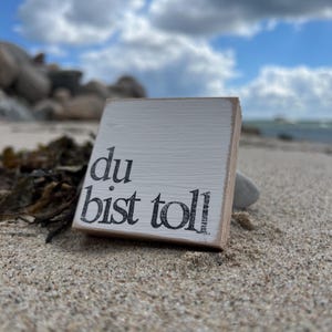May include: A white wooden sign with black lettering that reads "du bist toll" which translates to "you are great" in English. The sign is sitting on a sandy beach.