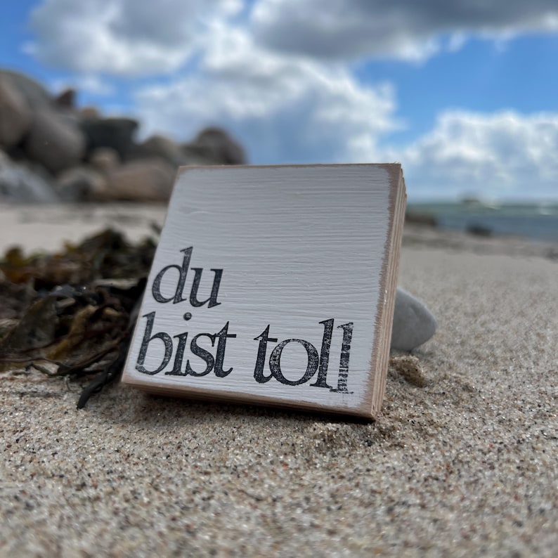May include: A white wooden sign with black lettering that reads "du bist toll" which translates to "you are great" in English. The sign is sitting on a sandy beach.