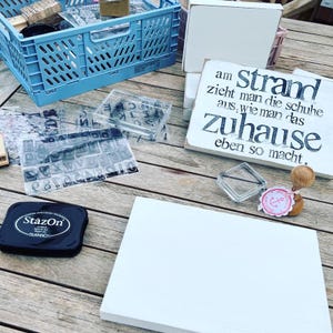 May include: A white wooden sign with black text that reads "am strand zieht man die schuhe aus, wie man das zuhause eben so macht."  The sign is sitting on a wooden table with other crafting supplies, including a black ink pad, clear stamps, and a small anchor stamp.