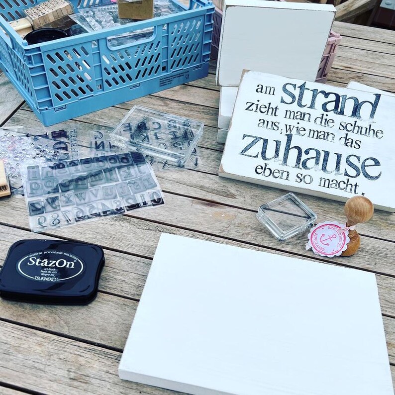 May include: A white wooden sign with black text that reads "am strand zieht man die schuhe aus, wie man das zuhause eben so macht."  The sign is sitting on a wooden table with other crafting supplies, including a black ink pad, clear stamps, and a small anchor stamp.