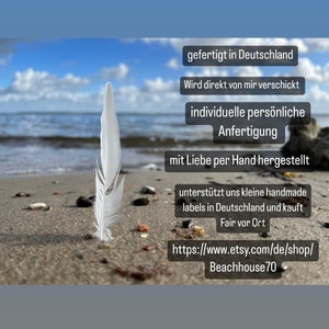 May include: A white feather lies on a sandy beach with the ocean in the background. The image includes text that reads "gefertigt in Deutschland", "Wird direkt von mir verschickt", "individuelle pers&ouml;nliche Anfertigung", "mit Liebe per Hand hergestellt", "unterst&uuml;tzt uns kleine handmade labels in Deutschland und kauft Fair vor Ort", and "https://www.etsy.com/de/shop/Beachhouse70".