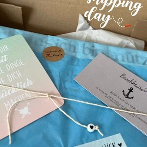 May include: A blue and white card with the text "Dir it e Dinge, e Dich ucklich machen" and a small heart with an arrow. A brown sticker with the text "handmade with love" is on the card. A white string with a black heart bead is tied around the card. A white card with the text "Beachhouse" and an anchor is in the background.