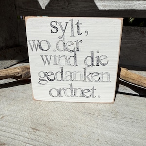 Könnte beinhalten: Ein kleines, rechteckiges Holzschild mit dem Text "Sylt, wo der Wind die Gedanken ordnet" in schwarzer Schrift. Das Schild hat eine verwitterte, cremefarbene Lackierung und ist wahrscheinlich ein Dekorationsartikel.