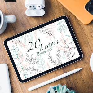 May include: A digital art setup on a wooden surface. A tablet displays a "20 Leaves Brush set" design. Accessories include a white stylus, AirPods, an orange phone, a succulent, and a coffee cup. The Procreate app icon is visible.