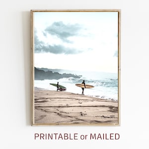 May include: Two surfers walk along a sandy beach with their surfboards. The ocean is in the background with waves crashing on the shore. The sky is cloudy and gray.
