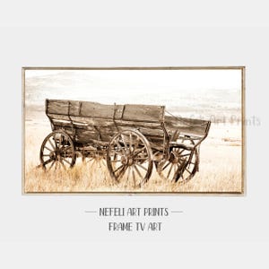 May include: Sepia-toned framed print featuring a weathered wooden wagon with large spoked wheels. The wagon sits in a field of dry grass, with a hazy mountain range in the background. The print is labeled "Nefeli Art Prints - Frame TV Art."