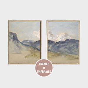 May include: Two rectangular wall art prints featuring landscape paintings of blue mountains under a pale sky with soft clouds. Each print is enclosed in a thin wooden frame. A circular badge at the bottom displays the text FRAMED or UNFRAMED.