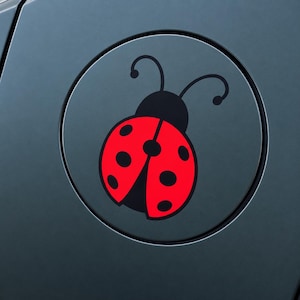 May include: A circular sticker featuring a graphic illustration of a ladybug with a bright red body and black spots. The ladybug has a black head and two curved antennae. The design is centered on a dark gray surface with a circular border.