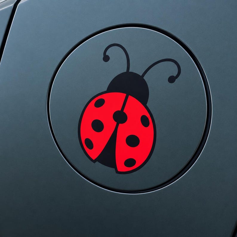 May include: A circular sticker featuring a graphic illustration of a ladybug with a bright red body and black spots. The ladybug has a black head and two curved antennae. The design is centered on a dark gray surface with a circular border.