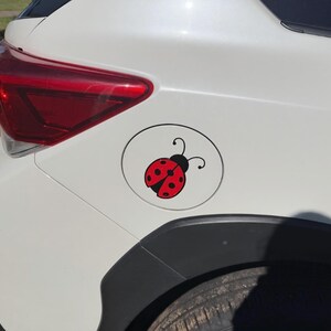 May include: A circular white car fuel door cover featuring a red and black ladybug decal. The ladybug has a bright red body with black spots and a black head with two antennae. The decal is centered on the fuel door of a white vehicle.