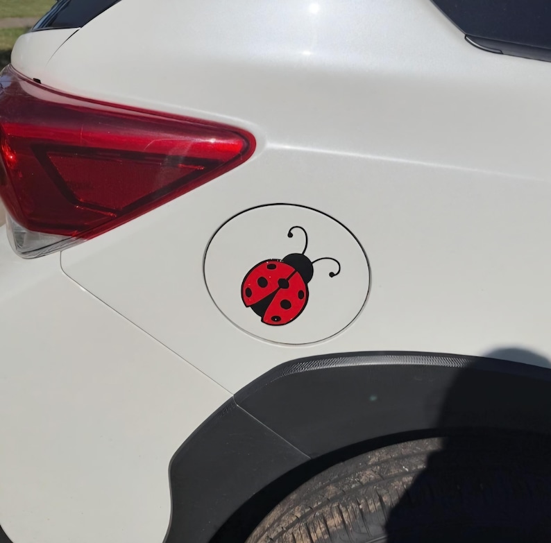 May include: A circular white car fuel door cover featuring a red and black ladybug decal. The ladybug has a bright red body with black spots and a black head with two antennae. The decal is centered on the fuel door of a white vehicle.