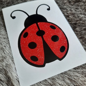 May include: A vinyl decal featuring a ladybug design on a white rectangular backing. The ladybug has a solid black head with two antennae and a red glitter body adorned with six distinct black circular spots and a central black stripe.