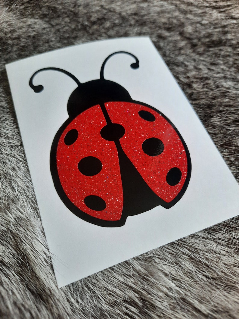 May include: A vinyl decal featuring a ladybug design on a white rectangular backing. The ladybug has a solid black head with two antennae and a red glitter body adorned with six distinct black circular spots and a central black stripe.