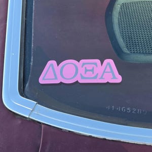 May include: A pink decal featuring the Greek letters Delta, Omicron, and Theta in a light gray color. The sticker is applied to a dark-tinted glass surface, displaying the characters in a bold, sans-serif font with a solid pink background.