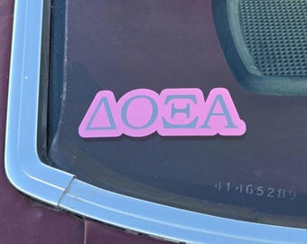 May include: A pink decal featuring the Greek letters Delta, Omicron, and Theta in a light gray color. The sticker is applied to a dark-tinted glass surface, displaying the characters in a bold, sans-serif font with a solid pink background.