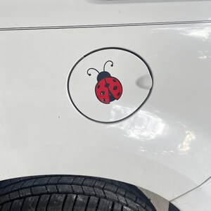 May include: A circular white fuel door cover on a white vehicle featuring a graphic decal of a red ladybug with black spots and black antennae. The ladybug is centered on the fuel door, which is positioned above a black tire on the vehicle.
