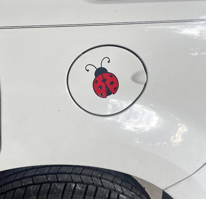 May include: A circular white fuel door cover on a white vehicle featuring a graphic decal of a red ladybug with black spots and black antennae. The ladybug is centered on the fuel door, which is positioned above a black tire on the vehicle.