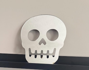Op de afbeelding: Zilverkleurige schedelvormige decoratie met uitgesneden oogkassen, een neusholte en een rij tanden. De schedel heeft een textuur en wordt getoond tegen een neutraal gekleurde muur en een zwarte plank.
