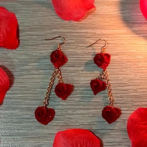 May include: A pair of heart-shaped dangle earrings. Each earring features three red heart charms suspended from a gold-colored chain. The earrings are displayed with red rose petals and lit candles.
