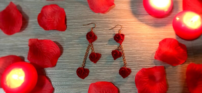 May include: A pair of heart-shaped dangle earrings. Each earring features three red heart charms suspended from a gold-colored chain. The earrings are displayed with red rose petals and lit candles.
