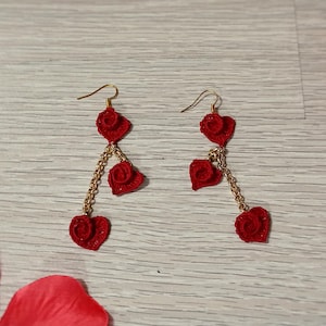 May include: A pair of red heart-shaped earrings with gold-colored chains and hooks. The earrings are made of a textured material and are displayed on a light wood-grain surface. Red rose petals and a red sphere are also visible.