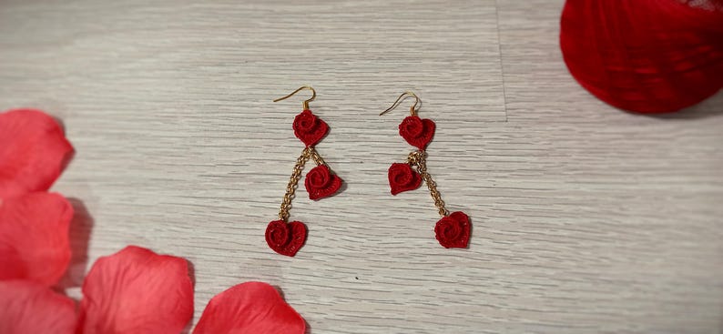 May include: A pair of red heart-shaped earrings with gold-colored chains and hooks. The earrings are made of a textured material and are displayed on a light wood-grain surface. Red rose petals and a red sphere are also visible.