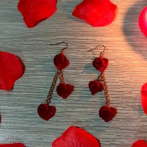 May include: A pair of heart-shaped earrings with a gold chain, surrounded by red rose petals and lit candles. The earrings have three heart charms dangling from the chain. The background is a light gray wooden surface.