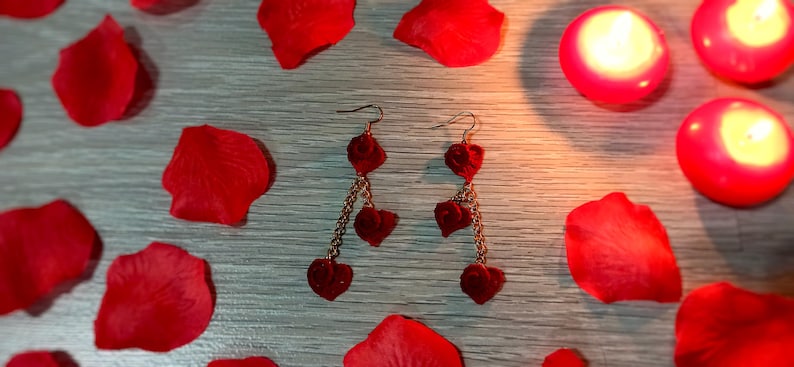 May include: A pair of heart-shaped earrings with a gold chain, surrounded by red rose petals and lit candles. The earrings have three heart charms dangling from the chain. The background is a light gray wooden surface.