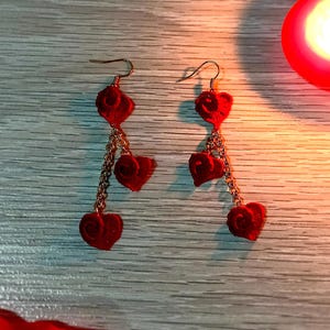 May include: A pair of red heart-shaped earrings with gold-colored chains. The earrings are dangling and have a velvet-like texture. The background includes red rose petals and lit candles, creating a romantic ambiance.