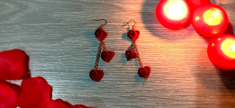 May include: A pair of red heart-shaped earrings with gold-colored chains. The earrings are dangling and have a velvet-like texture. The background includes red rose petals and lit candles, creating a romantic ambiance.