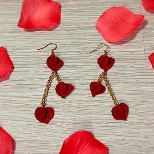 May include: A pair of red heart-shaped earrings with gold-colored chains and hooks, resting on a light wooden surface scattered with red rose petals. The earrings dangle with three heart charms.