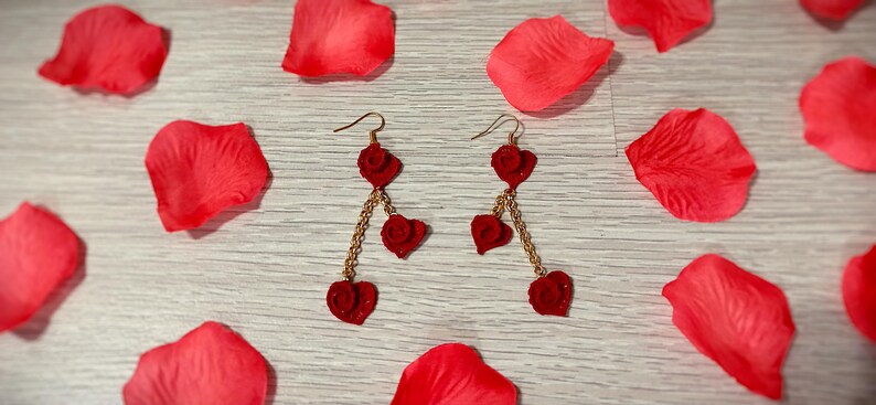 May include: A pair of red heart-shaped earrings with gold-colored chains and hooks, resting on a light wooden surface scattered with red rose petals. The earrings dangle with three heart charms.