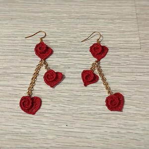 May include: A pair of red crochet heart earrings with gold-colored chain accents. Each earring features three heart shapes, with a small red ball at the center of each heart. A ball of red yarn is in the background.