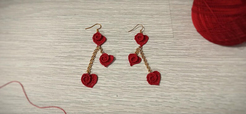 May include: A pair of red crochet heart earrings with gold-colored chain accents. Each earring features three heart shapes, with a small red ball at the center of each heart. A ball of red yarn is in the background.