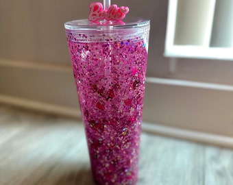 May include: A pink glitter tumbler with a clear lid and a pink straw. The tumbler has a pink "Barbie" charm on the lid.