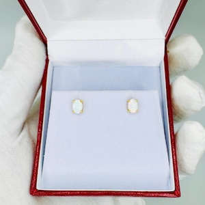 May include: A pair of oval-shaped opal stud earrings with gold settings, presented in a red jewelry box with a white interior. The earrings are displayed against a white background, showcasing their iridescent quality.