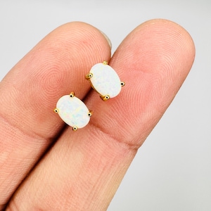 May include: A pair of oval-shaped opal stud earrings with gold-colored settings. The opals have a white base with iridescent flashes of color. The earrings are set with four small gold prongs.