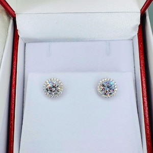 May include: A pair of round, sparkling stud earrings with a halo of smaller stones, presented in a red and white jewelry box. The earrings feature a central, clear stone surrounded by a ring of smaller, clear stones.