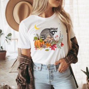 Peut inclure: T-shirt blanc avec une illustration à l'aquarelle d'un raton laveur entouré d'éléments automnaux. Le motif comprend une citrouille, des champignons et du feuillage d'automne. Un croissant de lune et un petit insecte sont également présents.