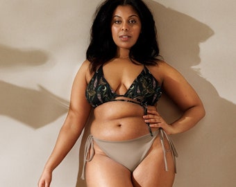 May include: A woman wearing a black and green patterned bikini top and a light brown bikini bottom with string ties. The bikini bottom has a low rise and is cut high on the legs.