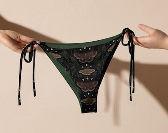 Op de afbeelding: Zwarte bikinibroek met een donkergroene rand en strikjes. De stof heeft een herhalend patroon van motten, manen en bladeren. De bikinibroek wordt door twee handen omhoog gehouden.