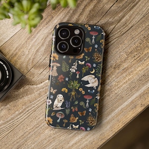 May include: A dark phone case with a whimsical forest pattern featuring owls, mushrooms, butterflies, and flowers. The case is on a wooden surface, showcasing the detailed design and vibrant colors.