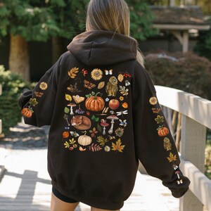 Puede incluir: Una sudadera con capucha negra con un gran estampado de elementos oto&ntilde;ales en la espalda. El estampado incluye una calabaza, setas, hojas, un zorro y una lib&eacute;lula.