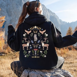 Peut inclure: Sweat à capuche noir avec un motif sur le thème de la nature au dos. Le motif représente un loup, des cerfs, des papillons et des éléments floraux. Le sweat à capuche est porté à l'extérieur avec un fond de montagne.