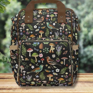 Op de afbeelding: Een zwarte rugzak met een bruin leren handvat en ritsen. De rugzak heeft een bloemenprint met paddenstoelen, vogels en bladeren.