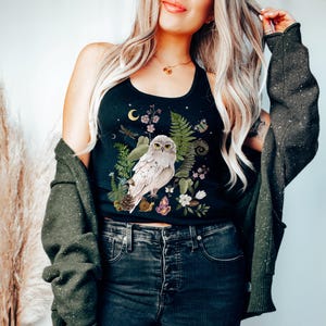 May include: Black tank top with a detailed illustration of a white owl surrounded by flowers, ferns, and crystals. The design includes a crescent moon and stars. The tank top is paired with dark wash jeans and a green cardigan.
