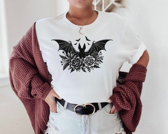 Könnte beinhalten: Weißes T-Shirt mit einem schwarzen Fledermaus-Design und floralen Elementen. Die Fledermaus hat ausgebreitete Flügel, umgeben von Rosen und Blättern. Das Outfit umfasst Jeansshorts, einen schwarzen Gürtel und eine bordeauxfarbene Strickjacke.