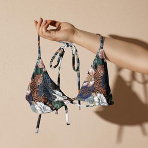 May include: A triangle-shaped bikini top with a vibrant tiger and floral print. The top features a dark blue background with orange tigers, pink and white flowers, and green foliage. The straps are dark blue with a matching pattern.