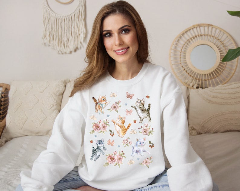 May include: White sweatshirt with a colorful print of kittens and butterflies. The kittens are in various poses, some are playing with yarn, others are sitting or standing. The butterflies are in flight and have a variety of colors.