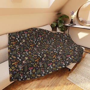May include: A dark blanket with a repeating pattern of mushrooms, ferns, insects, and other forest elements. The blanket is draped over a white couch. The design features a variety of colors, including red, brown, green, and yellow.